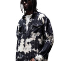 A man wearing a black and white tie-dye button-up shirt over a white t-shirt, styled with dark pants and sunglasses, standing in a modern concrete architectural setting.