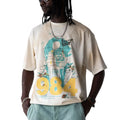 A man wearing a cream-colored graphic t-shirt with a blue and yellow '984' design, styled with light blue cargo shorts, white sunglasses, and a silver chain necklace against a light gray studio background.