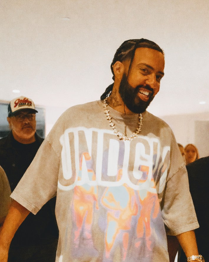 French Montana wearing Undergold with colorful graphics and text, standing indoors.