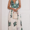 A woman modeling a sleeveless maxi dress with a green tropical palm leaf print on a cream background, holding a small beige handbag against a white paneled wall.