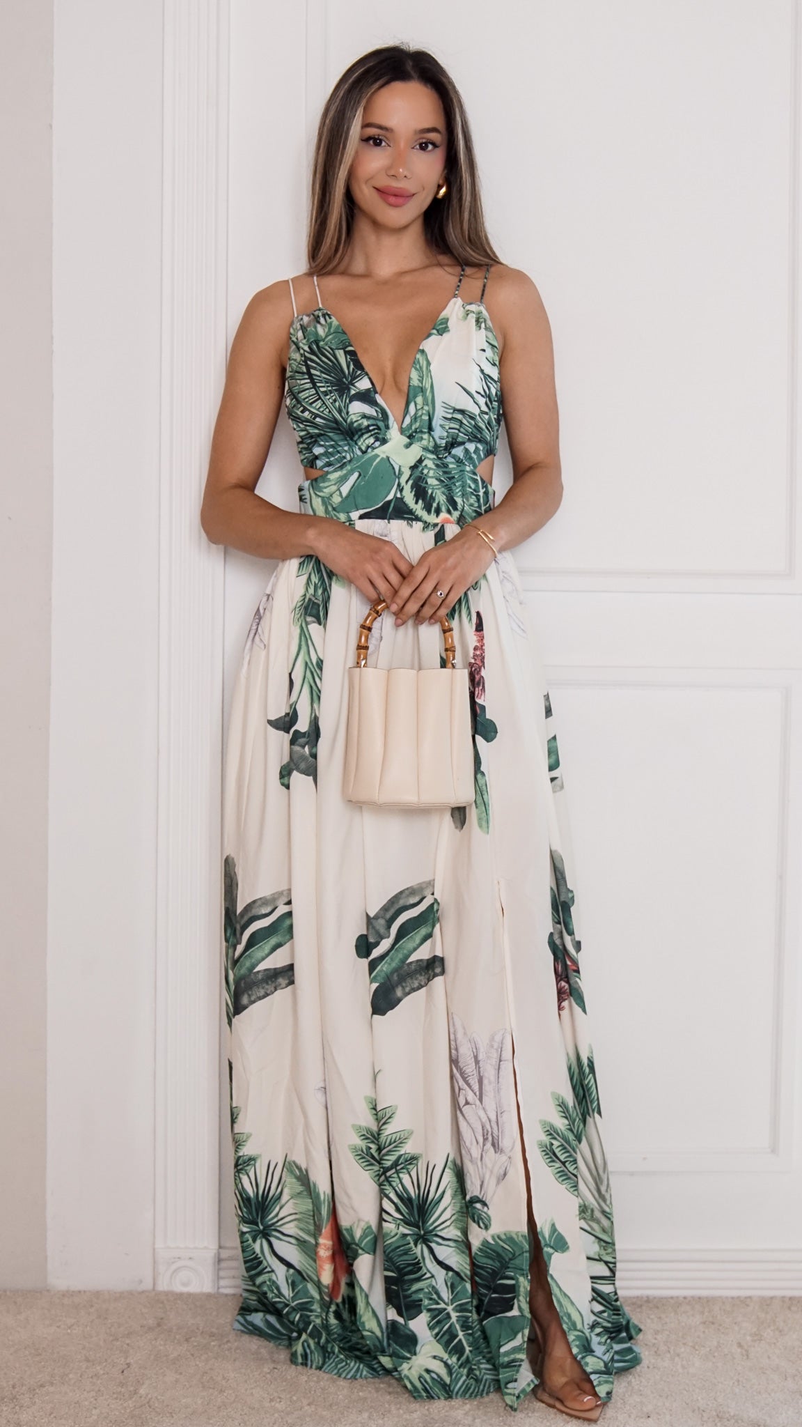 A woman modeling a sleeveless maxi dress with a green tropical palm leaf print on a cream background, holding a small beige handbag against a white paneled wall.