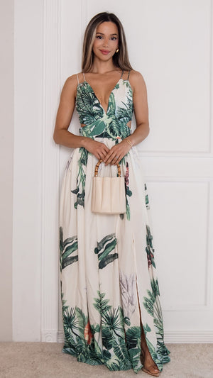 A woman modeling a sleeveless maxi dress with a green tropical palm leaf print on a cream background, holding a small beige handbag against a white paneled wall.
