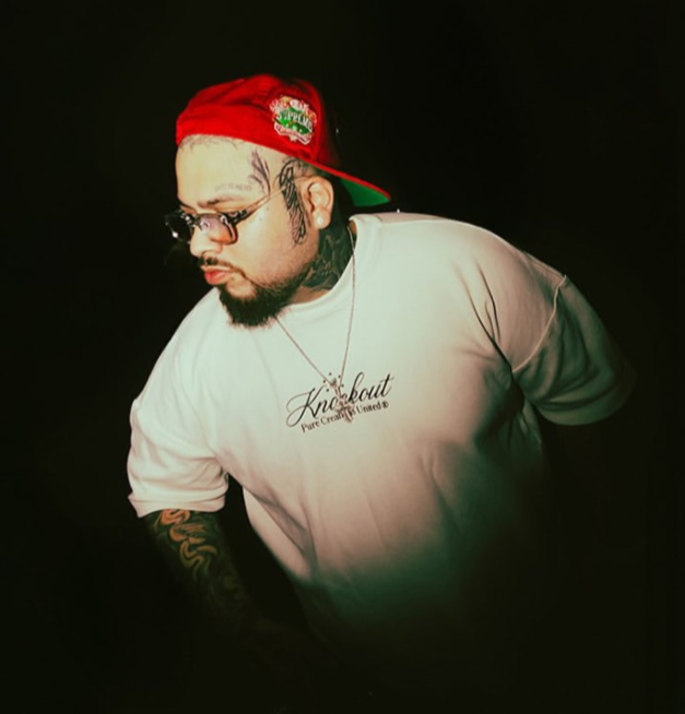 Totoy El Frio wearing a red cap and white t-shirt with text, against a dark background Pure Knockout