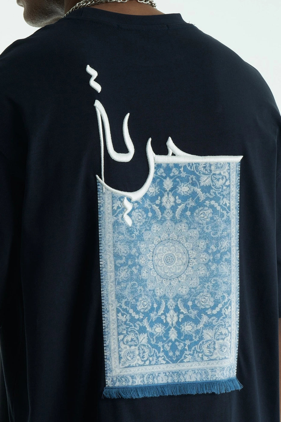 Black t-shirt with white Arabic calligraphy and a blue patterned rug graphic on the back